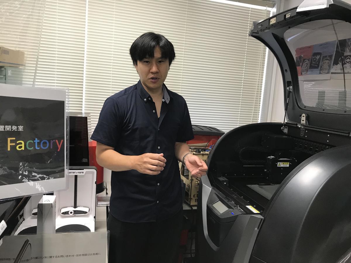 Engineer Ryusei Yamaguchi. On the right is the most popular type of 3D printer that is made in Japan, with its lid open. Also pictured are two low-priced Chinese printers, and one older model of American printer