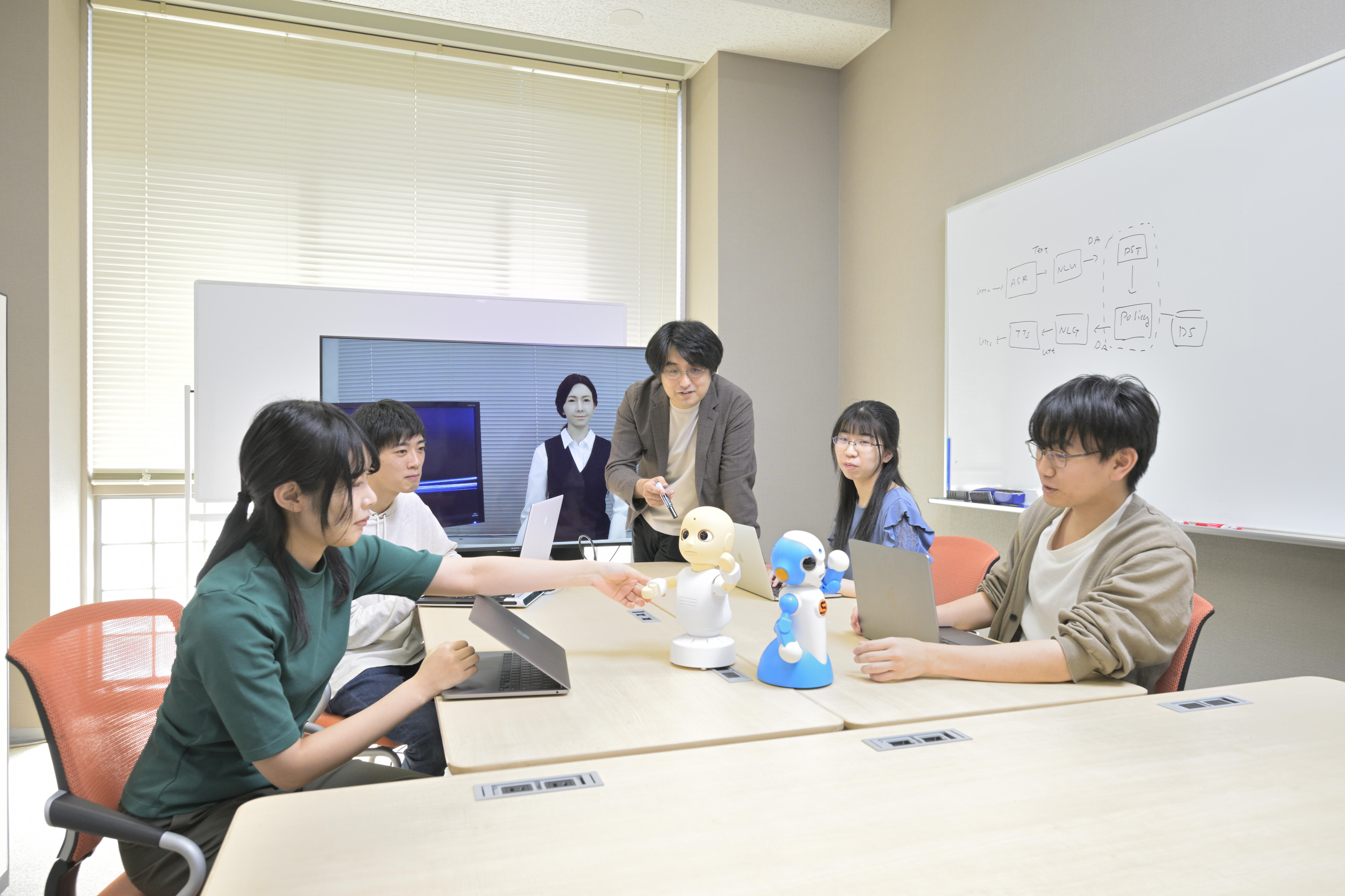 He and his students chat with the android Shoko-san (on screen) and the robots CommU (white) and SOTA (blue), which are used for researching dialogue systems.