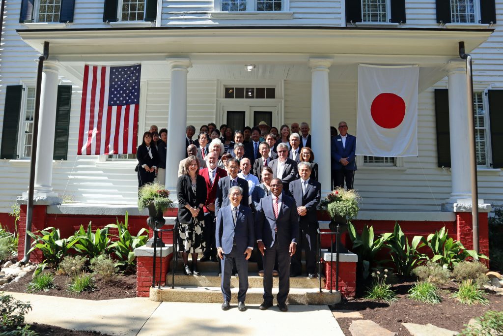The delegation from Nagoya University poses for a photo with the representatives from North Carolina State University at the entrance of a building, flanked by Japanese and American flags.