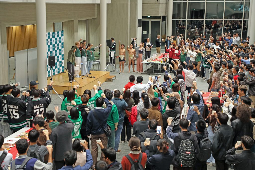 Students face the stage and kick off the party with a toast, shouting "kanpai!"