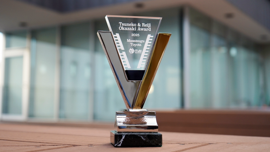 A photograph of the trophy for the 9th Tsuneko & Reiji Okazaki Award, resembling a 'V' with one side painted gold and the other silver. It reads "Tsuneko & Reiji Okazaki Award 2025, Masatsugu Toyota, ITbM Nagoya University."