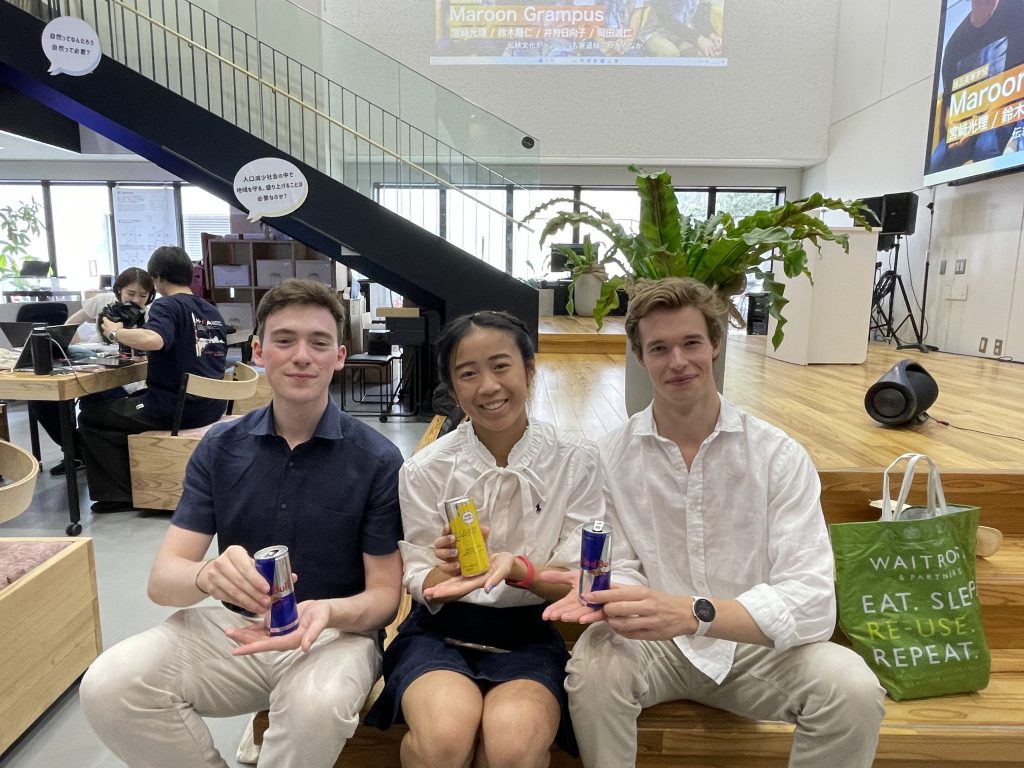 Three students from St John's College sit on the stage at Idea Stoa and hold up the free energy drinks they received.