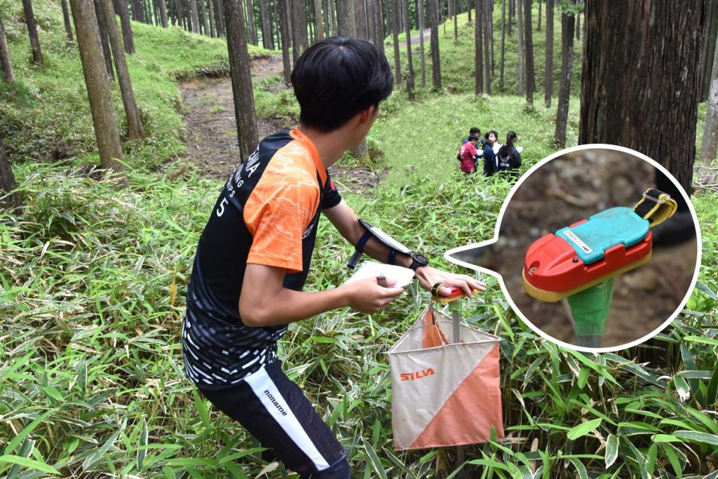 A student check in at a control using their unique tag while looking at a group of three students in the distance.