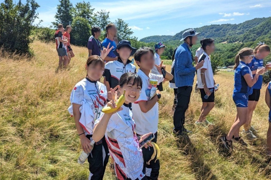 Four students from the Japan national team pose for a photograph at the 2023 Junior World Championship. Participants from other countries stand in the background, looking off in the distance.