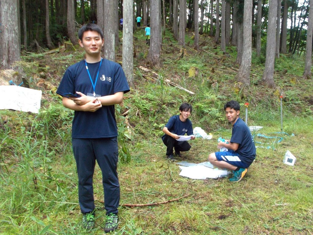 Two students crouch down to install a checkpoint in the forest, while another stands nearby facing the camera.