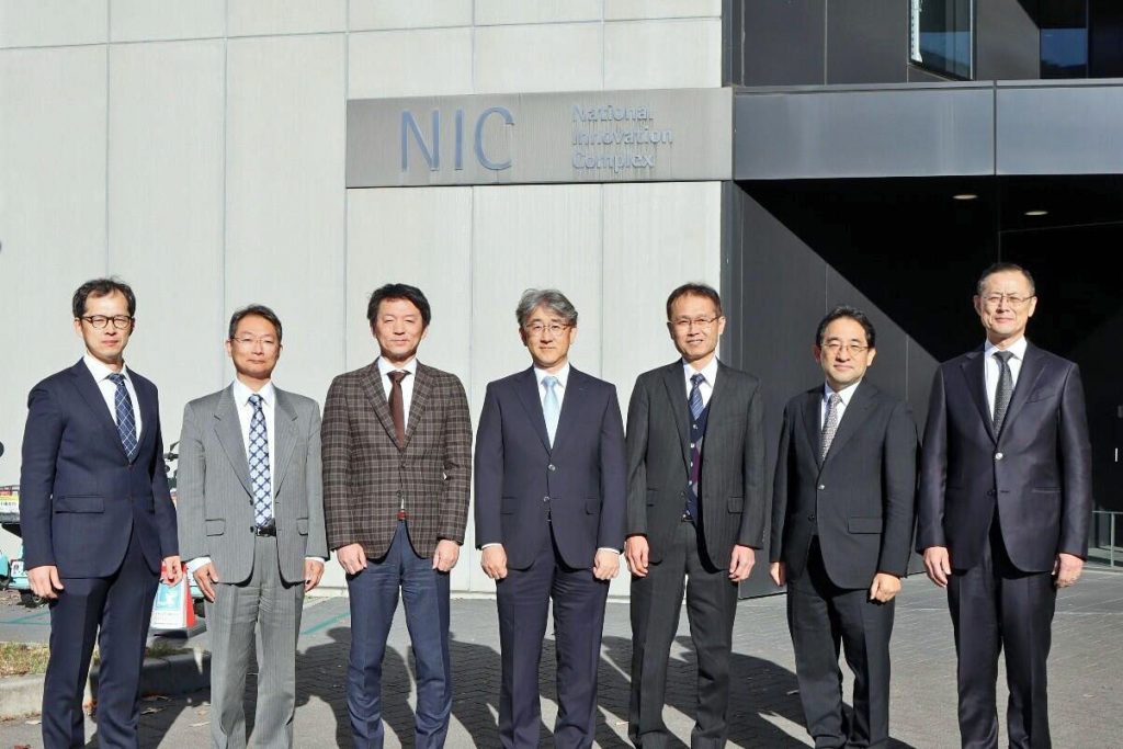 Seven men in suits from Brother Industries and Nagoya University stand in front of the building on campus that will house the new joint research laboratory.