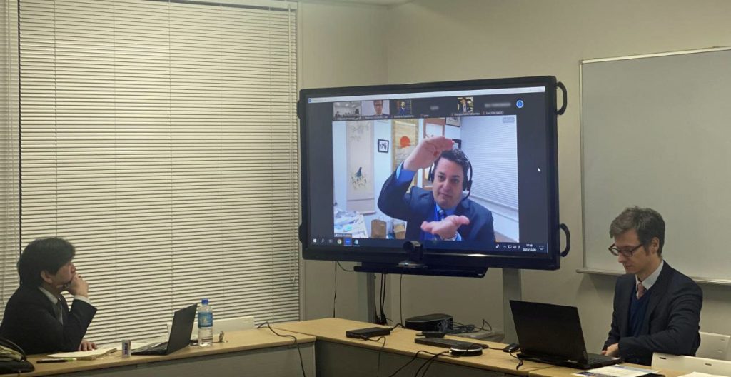 A scene from a pervious hybrid event; two men sit at a table while a third participates via Zoom.
