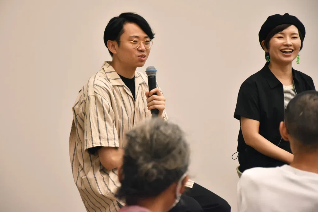 Sitting on a stool, Mizuno talks while holding a microphone during an event at Common Nexus.