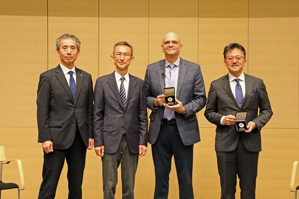 Four men stand in a line on stage; two of them hold the Nagoya Medals that they were awarded.