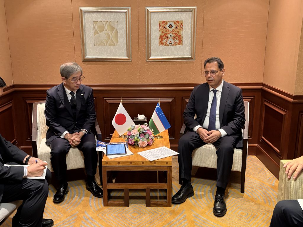 Two men sit on opposite sides of a low table, on which small Japanese and Uzbek flags are placed.