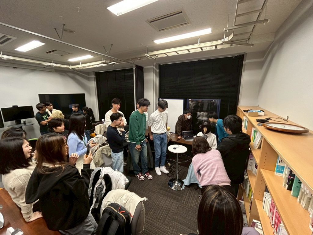 Students stand around in a room while learning about a project related to robotics.
