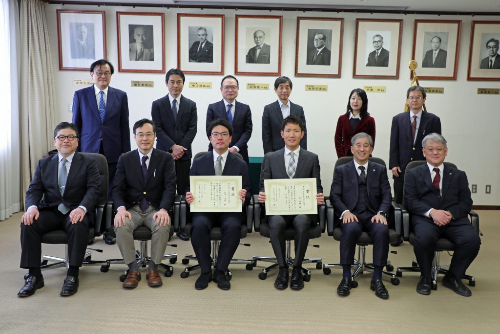 A row of six people stand behind a row of six people seated in chairs; the two recipients sit in the middle of the front row, holding their certificates.