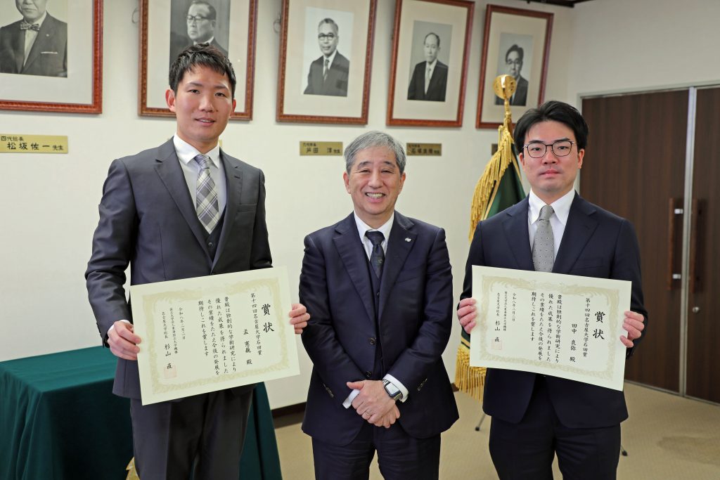 President Sugiyama stands in a row between the two recipients, who hold their certificates.