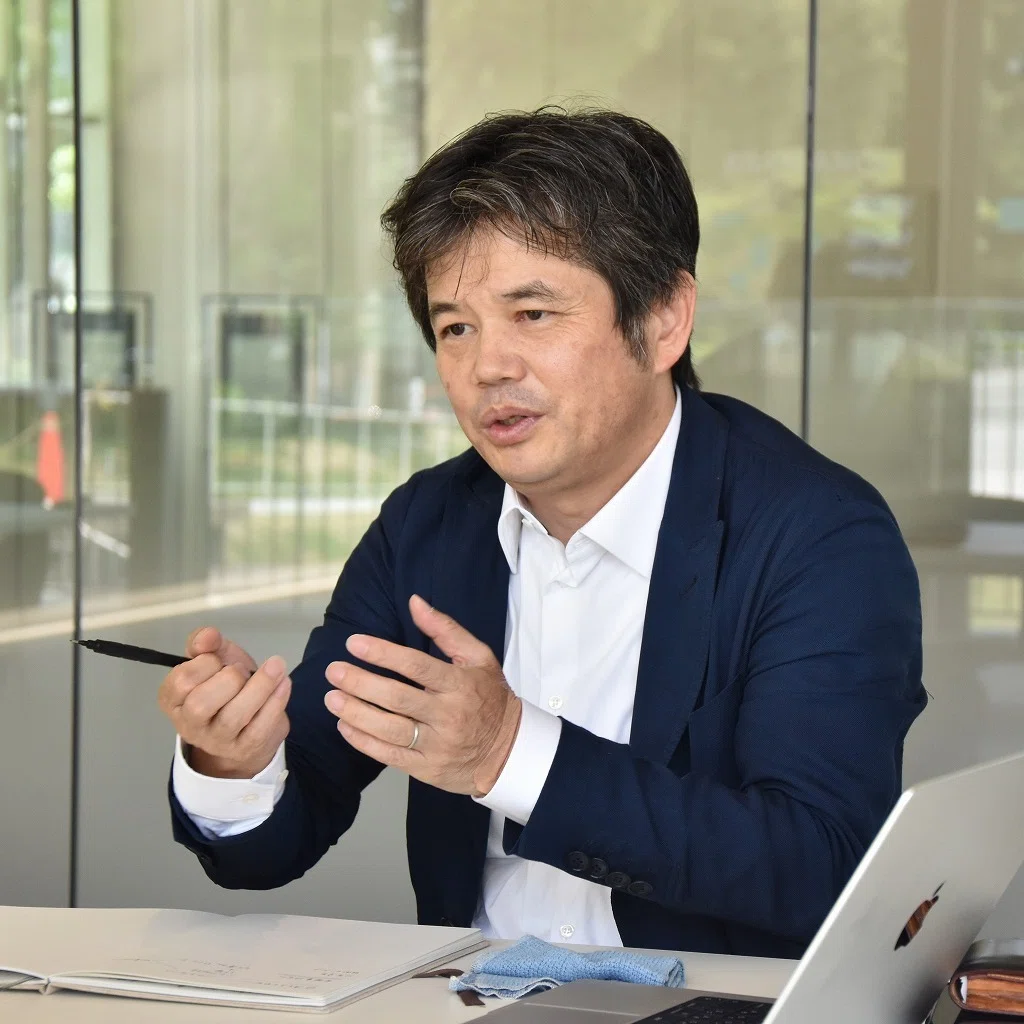 A man sits at a table, gesturing as he gives an explanation. He holds a pen in his right hand.