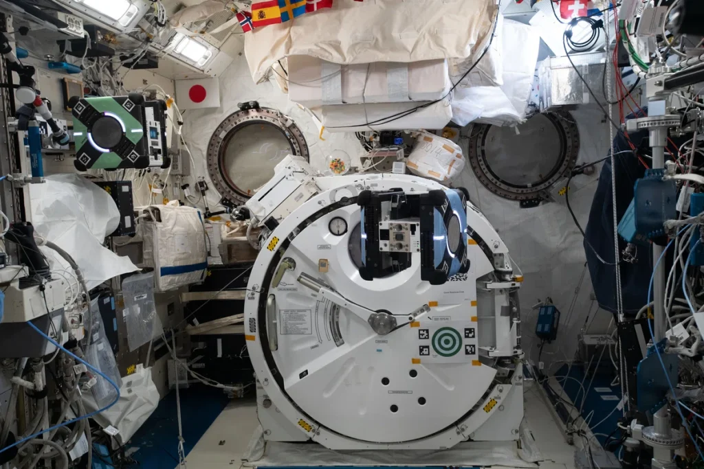 A photograph of the Japanese Experiment Module "Kibo" on the ISS. Two cube-shaped drones are pictured in flight, one green, the other blue.