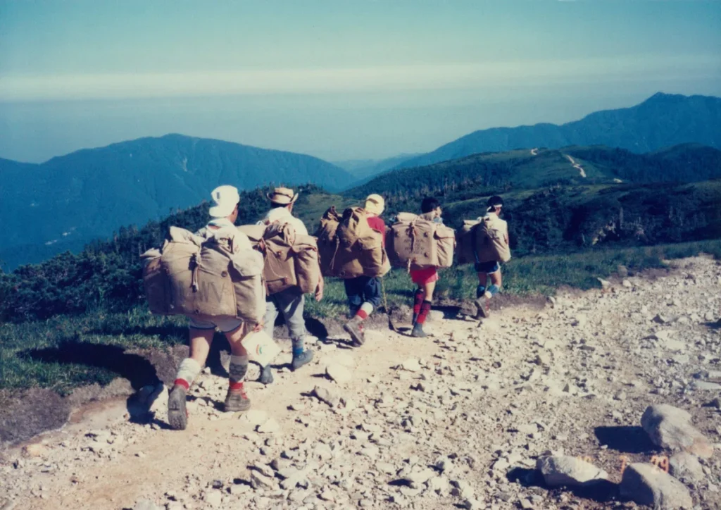 A film photograph shows five students walking along a mountain ridge, each carrying a large bag on their back.