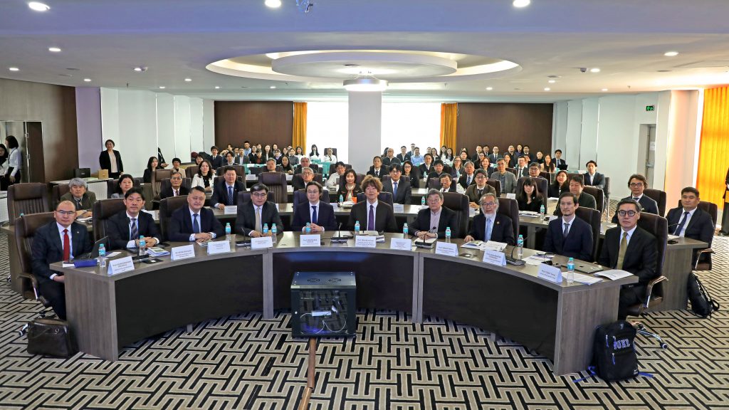 The anniversary symposium participants sit a semicircular tables for a commemorative group photo, taken from the stage at the front of the room.