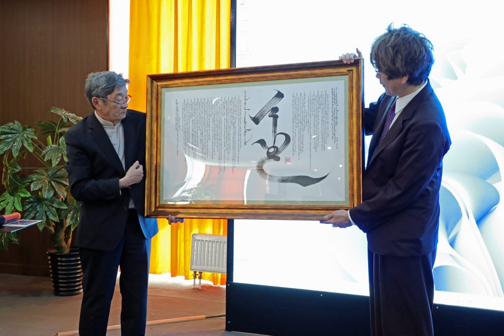 Professor Narangerel and Vice Dean Tamura face each other on stage, holding a large framed document.