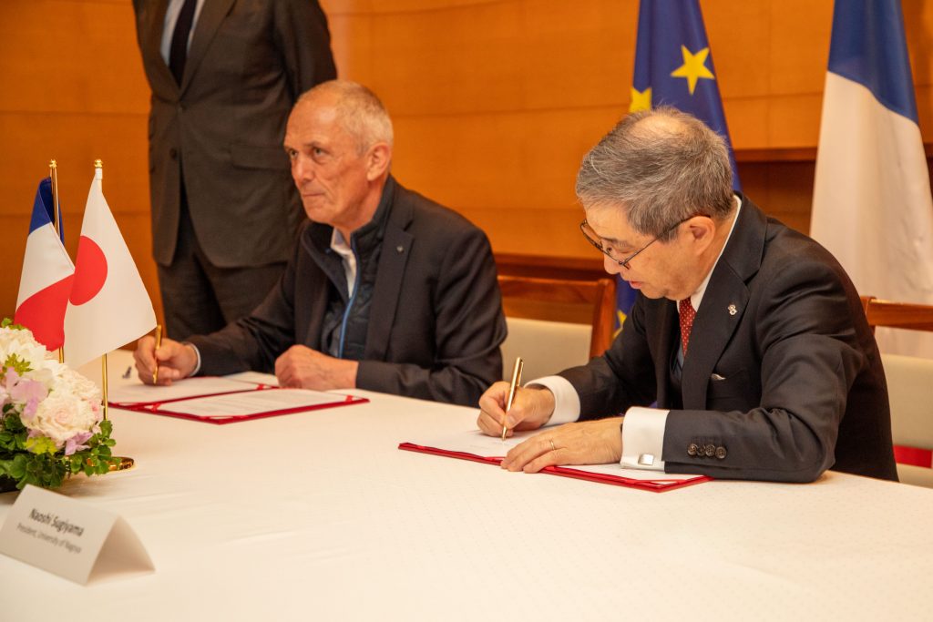Naoshi Sugiyama and Antoine Petit sit next to each other at a long table, each man signing a copy of the agreement.