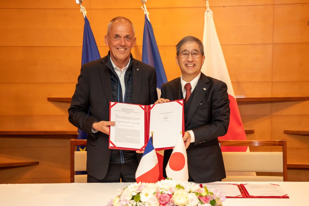 Naoshi Sugiyama and Antoine Petit stand, jointly holding up a signed copy of the agreement.