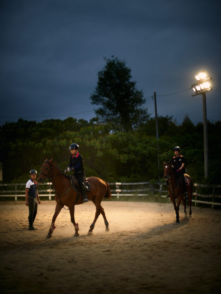 Two students on horseback in the dim light during evening training, accompanied by their senior.